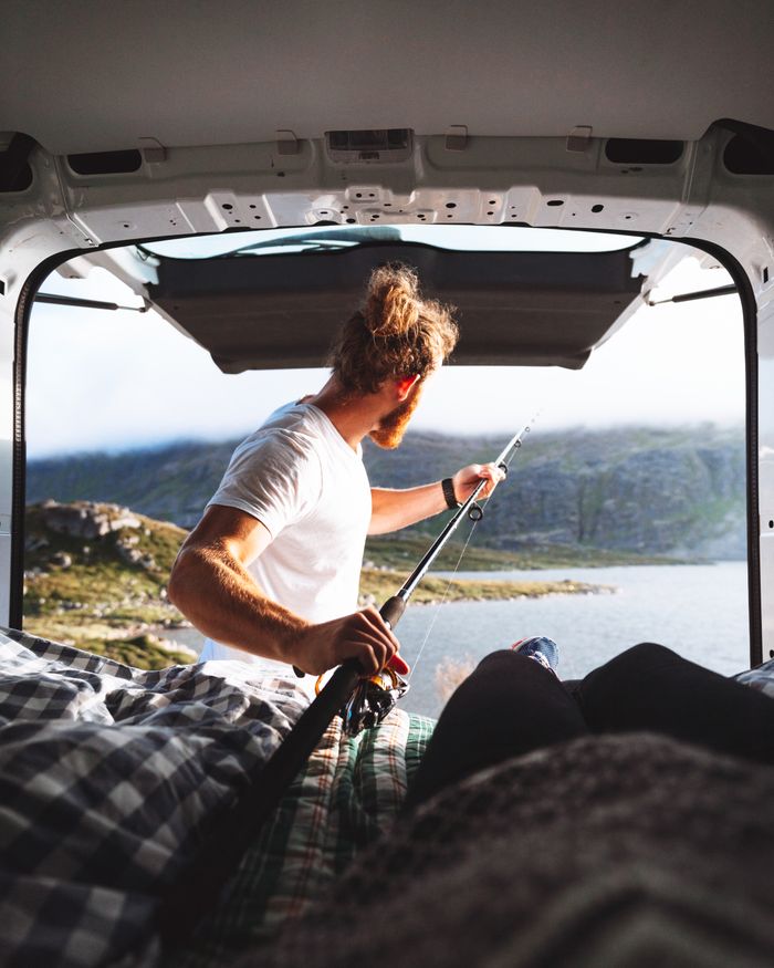 Man enjoys fishing from a van with a scenic lake view, embracing outdoor adventure and travel.
